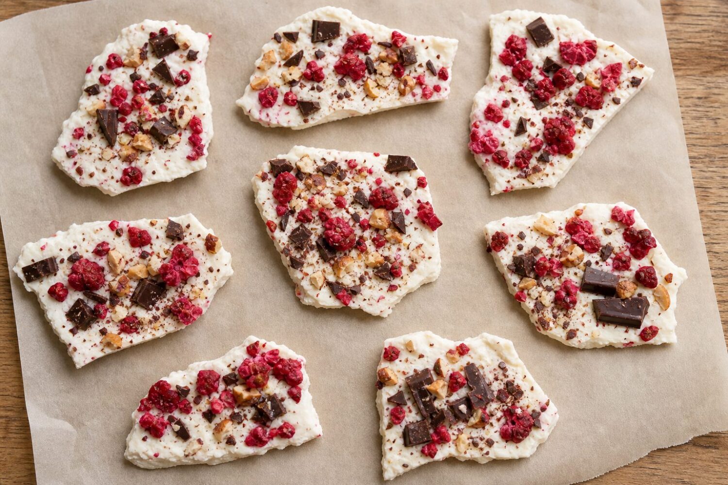Broken pieces of frozen yogurt bark scattered on parchment with freeze-dried raspberries and nuts
